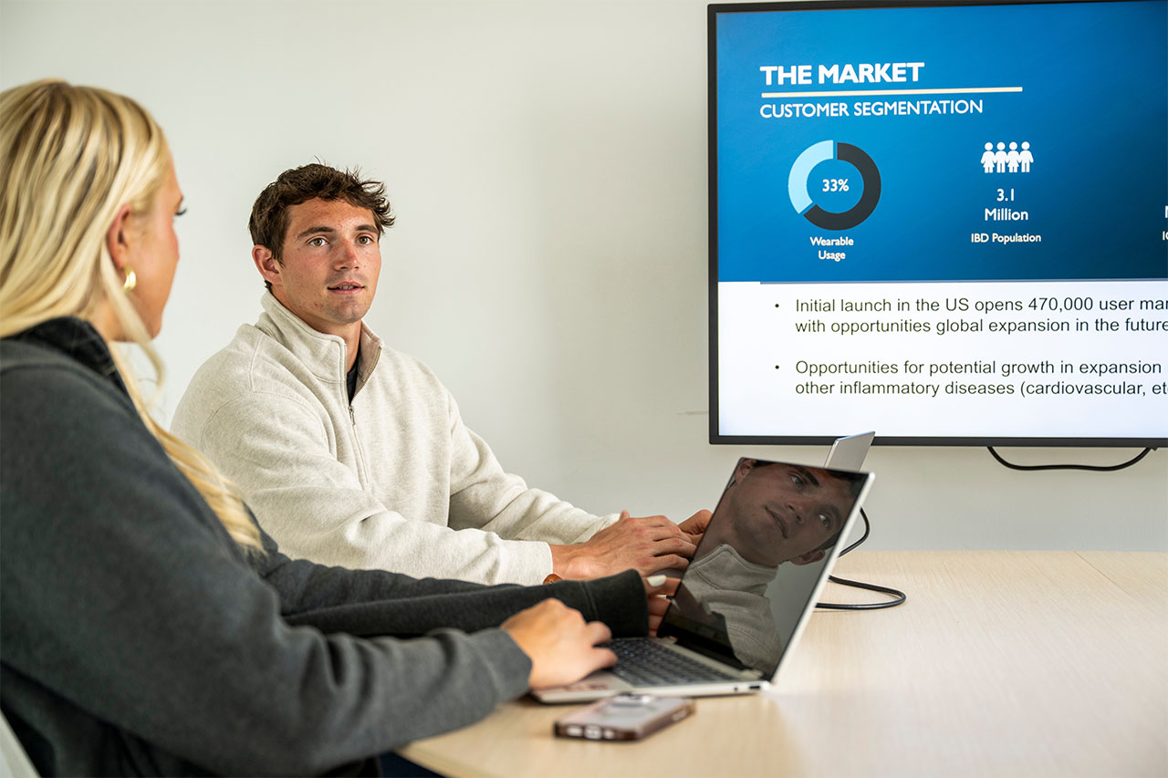 Two people sit in front of laptops on a conference table while a presentation is displayed on a monitor at the head of the table. The presentation has a heading that says 'The Market, Customer Segmentation'